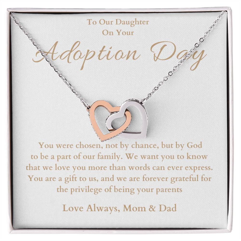 he image shows an open jewelry box with a necklace featuring two interlocking hearts, one silver and one rose gold. The background of the box has a message for adoption day to a girl