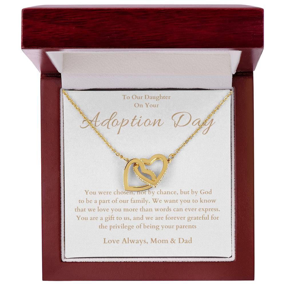 he image shows an open jewelry box with a necklace featuring two interlocking hearts, one silver and one rose gold. The background of the box has a message for adoption day to a girl