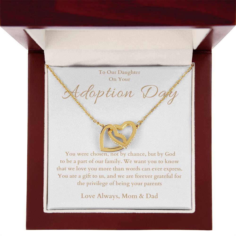 he image shows an open jewelry box with a necklace featuring two interlocking hearts, one silver and one rose gold. The background of the box has a message for adoption day to a girl