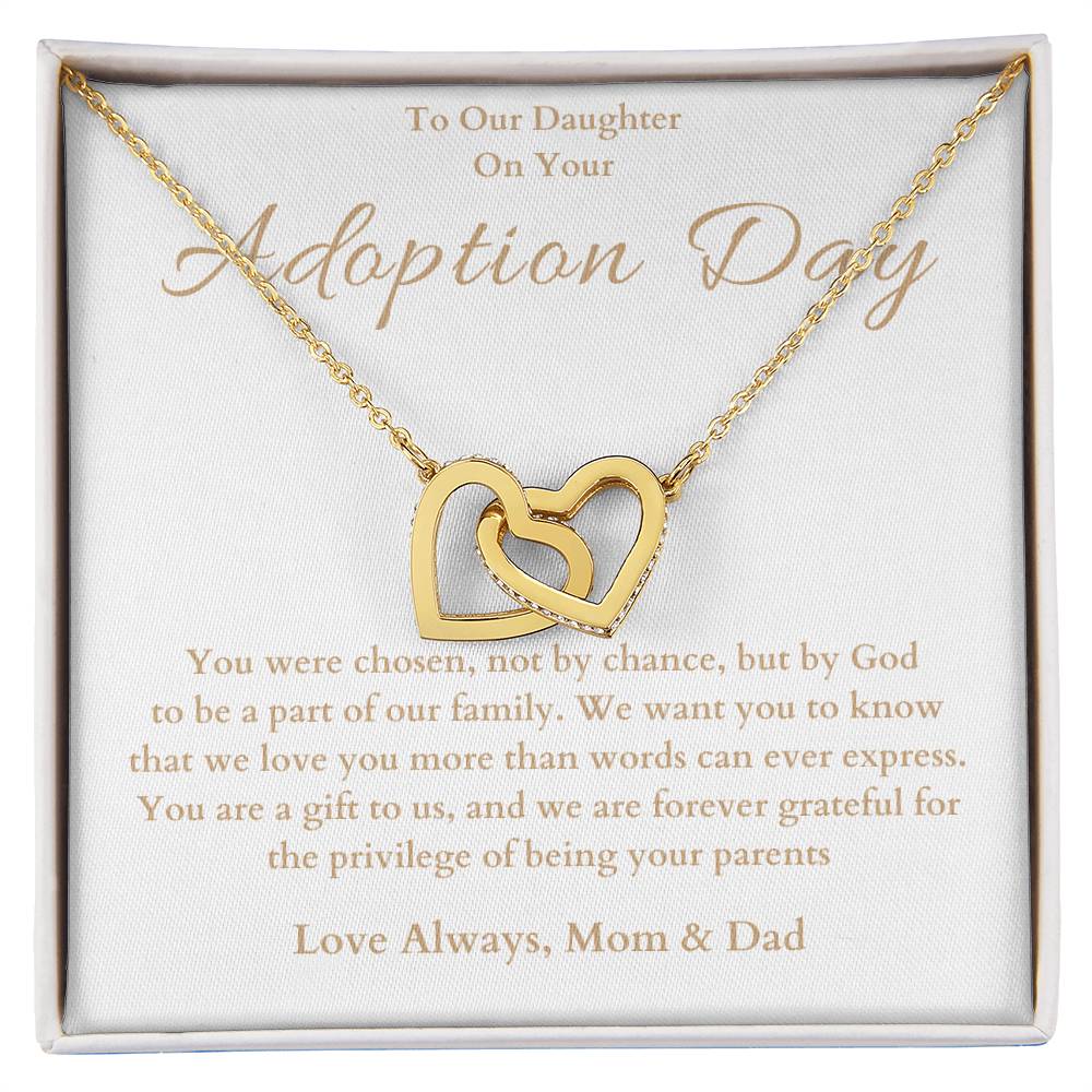 he image shows an open jewelry box with a necklace featuring two interlocking hearts, one silver and one rose gold. The background of the box has a message for adoption day to a girl