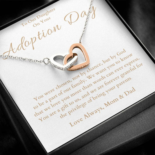 he image shows an open jewelry box with a necklace featuring two interlocking hearts, one silver and one rose gold. The background of the box has a message for adoption day to a girl