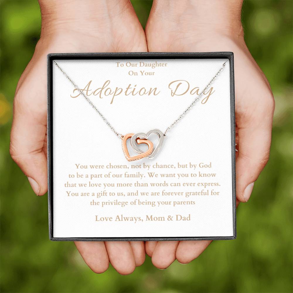 he image shows an open jewelry box with a necklace featuring two interlocking hearts, one silver and one rose gold. The background of the box has a message for adoption day to a girl