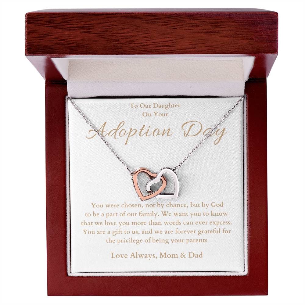 he image shows an open jewelry box with a necklace featuring two interlocking hearts, one silver and one rose gold. The background of the box has a message for adoption day to a girl